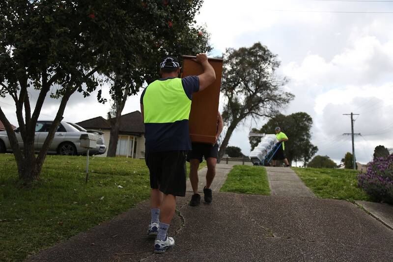 Two removal workers in high-visibility vests carrying a wooden cabinet from a suburban property