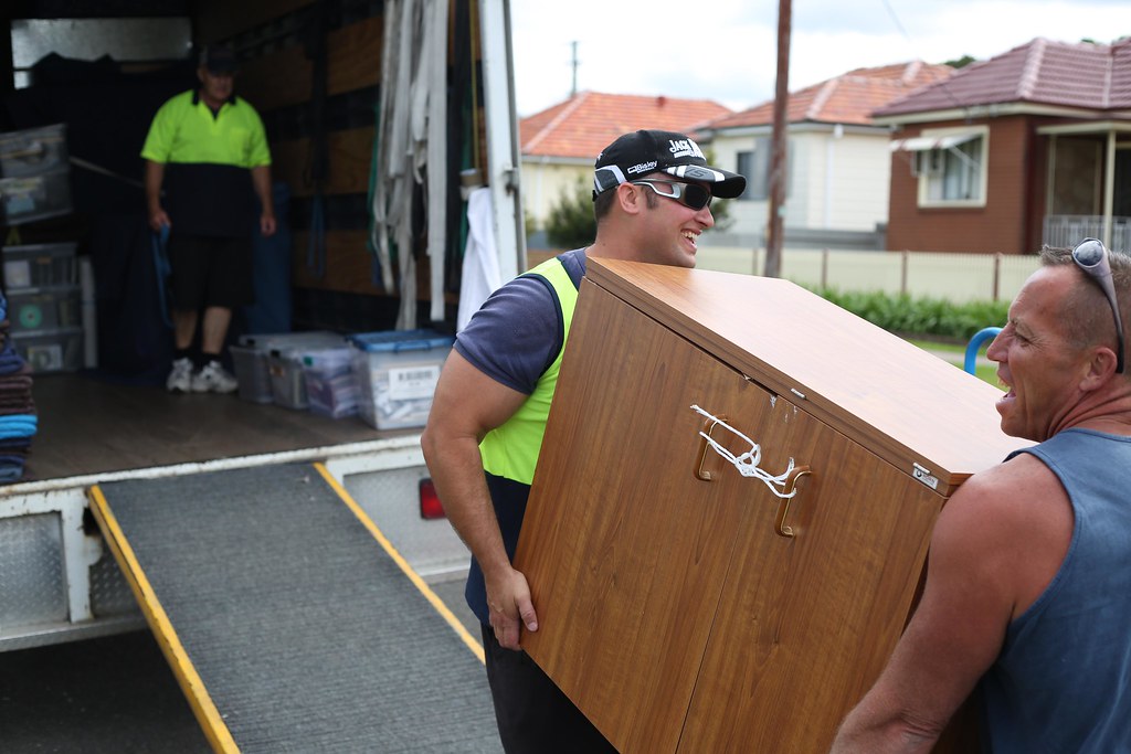 Two removal workers carrying a wooden cabinet away from a truck on a suburban street